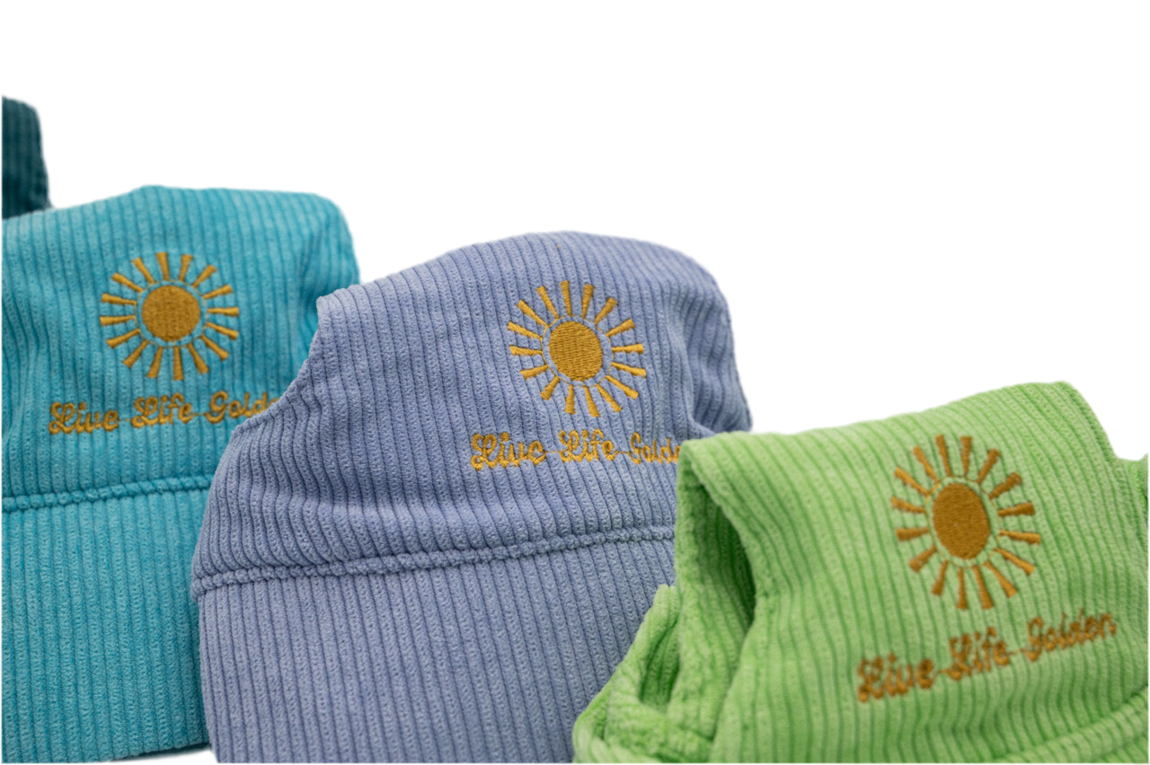 Close-up of embroidered sunburst with ‘Live Life Golden’ text on corduroy dog hats in purple, green, and teal.