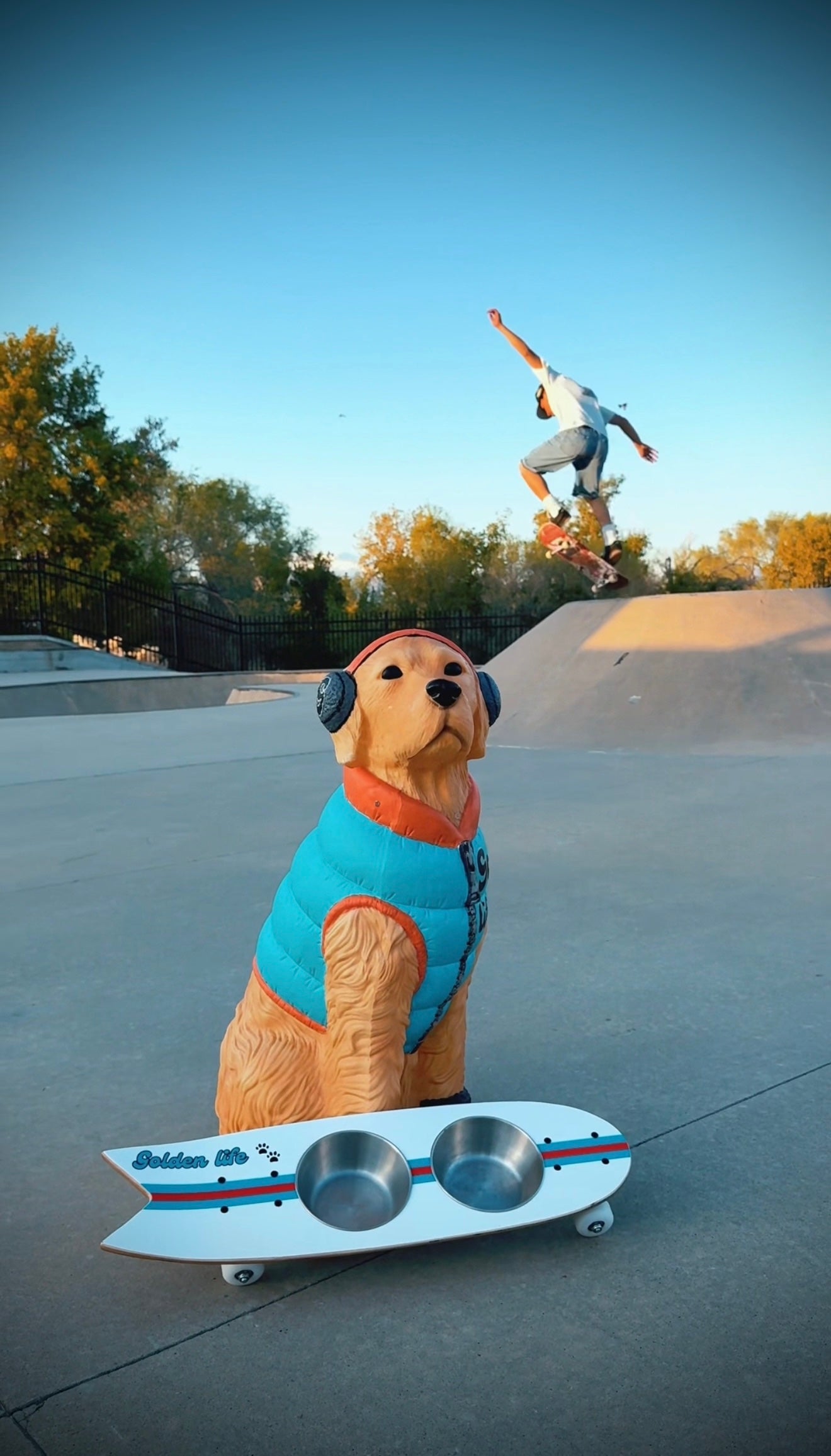 The Golden Life Kickflip Kibble Board dog feeder shaped like a skateboard with two stainless steel bowls, white board design with teal and orange racing stripes, and fixed black wheels. Get your Skateboard Dog Bowl today!