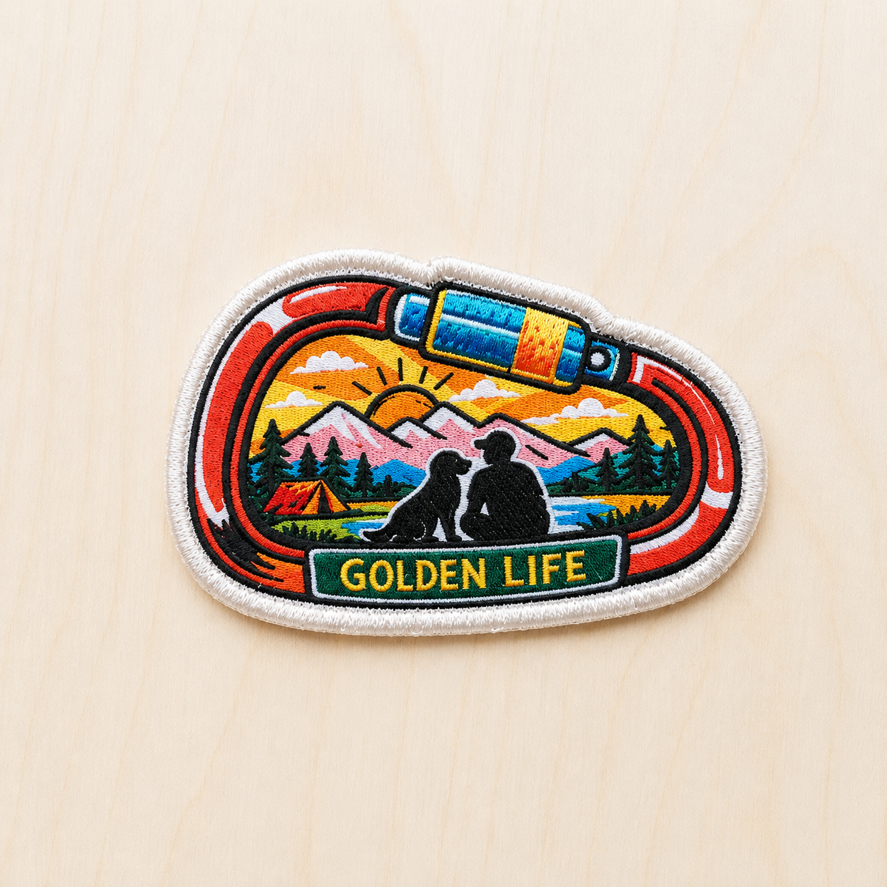 Adventure-themed embroidered iron-on patch shaped like a climbing carabiner featuring mountains, a sunset, pine trees, a camper tent, and a silhouette of a dog and person beside a lake with “Golden Life” text on a light wood background.