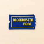 Embroidered retro Blockbuster video rental ticket-style iron-on patch inspired by classic movie rental stores, featuring blue and yellow stitched lettering on a light wood background.