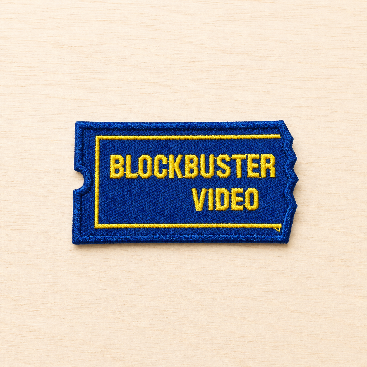 Embroidered retro Blockbuster video rental ticket-style iron-on patch inspired by classic movie rental stores, featuring blue and yellow stitched lettering on a light wood background.