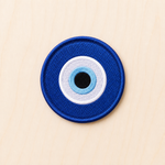Round embroidered blue evil eye iron-on patch featuring layered blue, white, and black concentric circles on a light wood background.