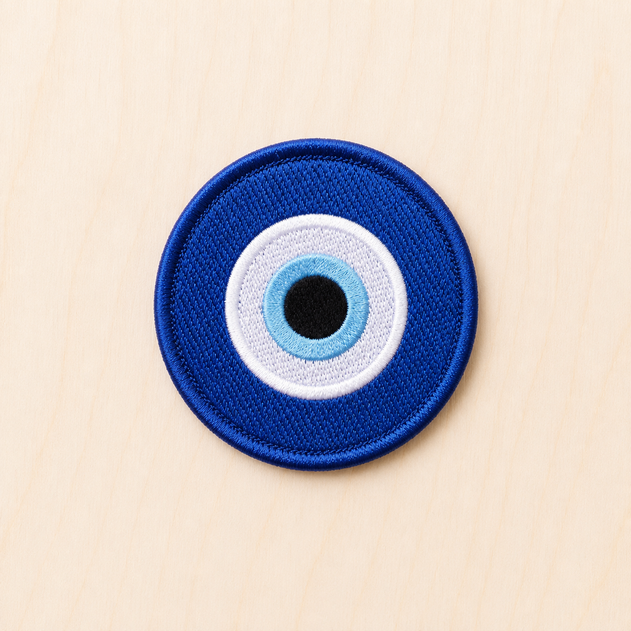 Round embroidered blue evil eye iron-on patch featuring layered blue, white, and black concentric circles on a light wood background.