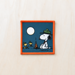 Retro-inspired embroidered patch featuring a cartoon-style white dog and two small bird companions wearing camping hats while sitting beside a campfire under a bright full moon, bordered with orange stitching and displayed on a light wood surface.
