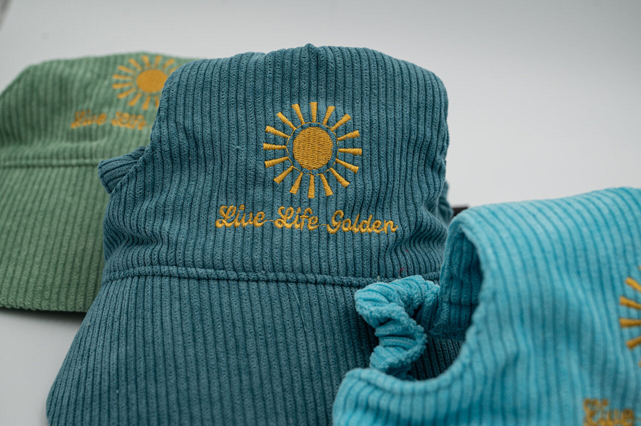 Close-up blue corduroy dog hat with golden sunburst and ‘Live Life Golden’ embroidery, with green and teal hats in background.