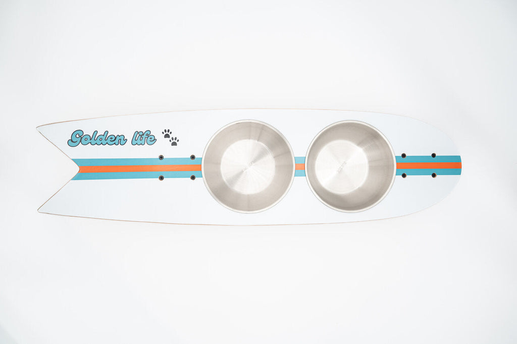 The Golden Life Kickflip Kibble Board dog feeder shaped like a skateboard with two stainless steel bowls, white board design with teal and orange racing stripes, and fixed black wheels. Get your Skateboard Dog Bowl today!