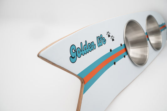 The Golden Life Kickflip Kibble Board dog feeder shaped like a skateboard with two stainless steel bowls, white board design with teal and orange racing stripes, and fixed black wheels. Get your Skateboard Dog Bowl today!