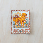 Stamp-style embroidered iron-on patch featuring a golden retriever standing in a wildflower field with retro sunset scenery and “FIRST CLASS PUP” text in warm vintage-inspired colors on a light wood background.