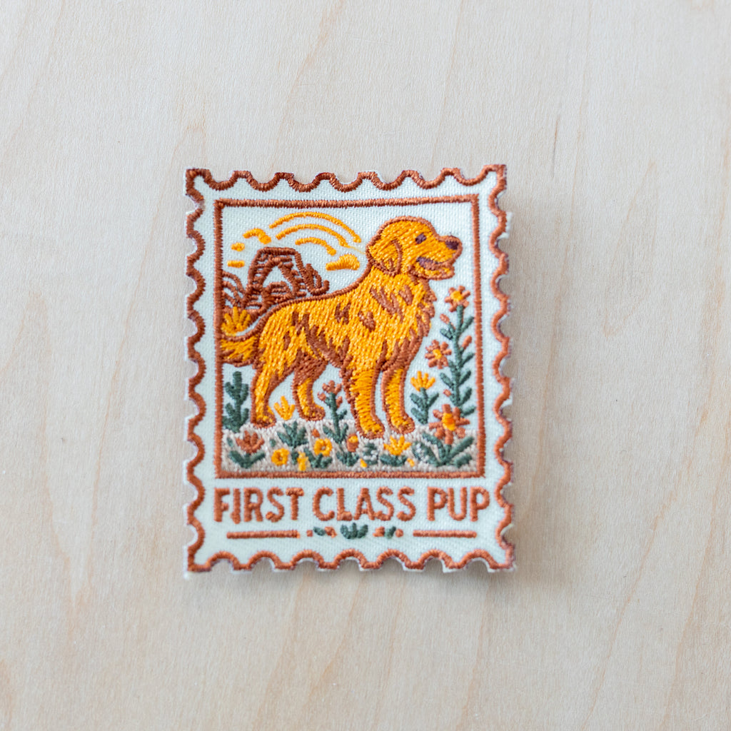 Stamp-style embroidered iron-on patch featuring a golden retriever standing in a wildflower field with retro sunset scenery and “FIRST CLASS PUP” text in warm vintage-inspired colors on a light wood background.