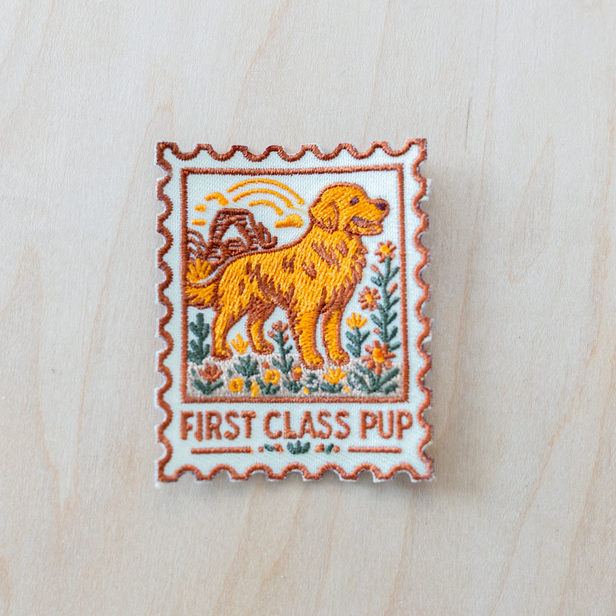 Stamp-style embroidered iron-on patch featuring a golden retriever standing in a wildflower field with retro sunset scenery and “FIRST CLASS PUP” text in warm vintage-inspired colors on a light wood background.