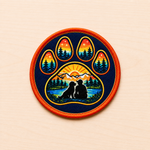 Round embroidered paw print iron-on patch featuring a sunset mountain landscape with pine trees, a dog and person silhouette beside a lake, and warm orange and blue retro outdoor colors on a light wood background.