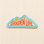 Mountain-shaped embroidered iron-on patch featuring “Golden Life” text with a rising sun design in orange, yellow, and blue on a light wood background.