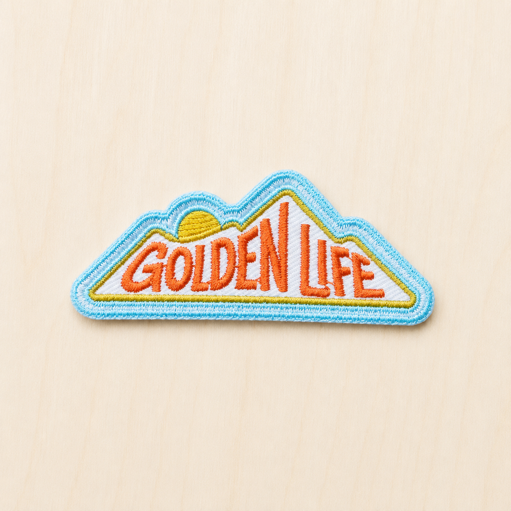 Mountain-shaped embroidered iron-on patch featuring “Golden Life” text with a rising sun design in orange, yellow, and blue on a light wood background.