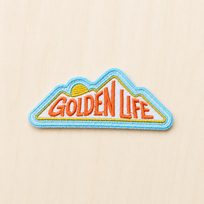 Mountain-shaped embroidered iron-on patch featuring “Golden Life” text with a rising sun design in orange, yellow, and blue on a light wood background.