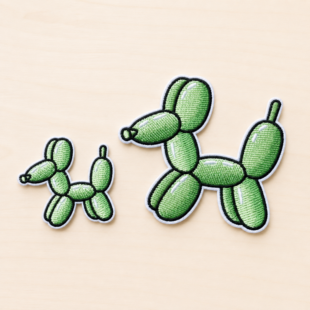 Set of two embroidered green balloon dog iron-on patches in small and large sizes with black and white stitched borders on a light wood background.