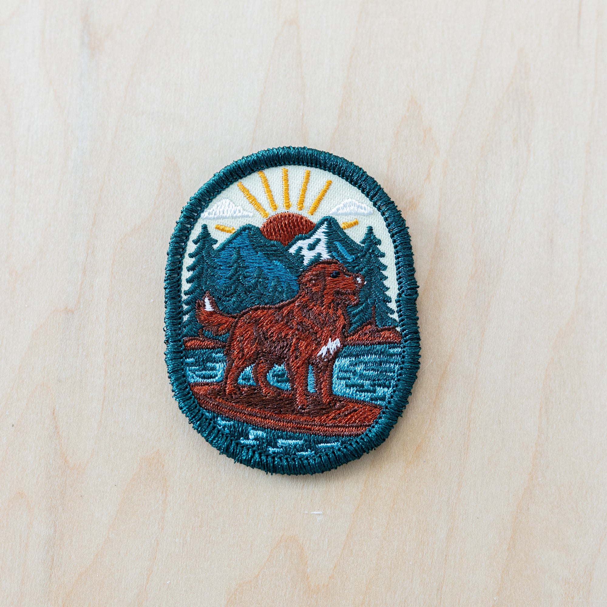 Oval embroidered patch featuring a brown adventure dog standing on a paddleboard in a mountain lake surrounded by pine trees and snowy peaks at sunrise, bordered with teal stitching and displayed on a light wood surface.