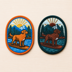 Side-by-side comparison of two oval embroidered paddleboarding dog patches featuring mountain lake scenes at sunrise. One patch shows a golden-colored adventure dog with bright blue and orange accents, while the newer version features a darker brown dog with deeper forest green and teal tones, both displayed on a light wood surface.