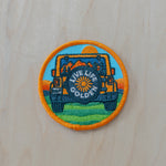 Round embroidered iron-on patch featuring an off-road Jeep with a “LIVE LIFE GOLDEN” spare tire cover, retro mountains, sunset, and outdoor scenery in bright orange, teal, blue, and green tones on a light wood background.