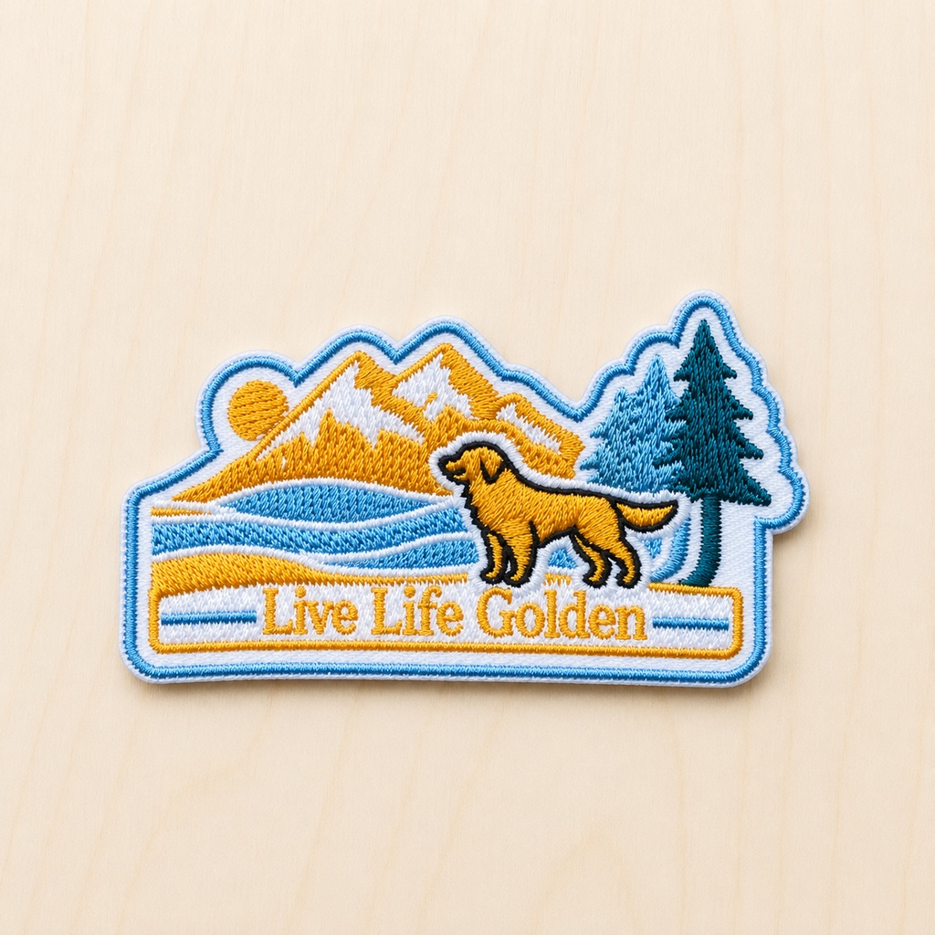Embroidered iron-on patch featuring a golden retriever in front of mountains, pine trees, and a lake with “Live Life Golden” text in a retro outdoor adventure style on a light wood background.