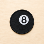 Round embroidered black magic 8 ball iron-on patch with a white circle and black number 8 design on a light wood background.