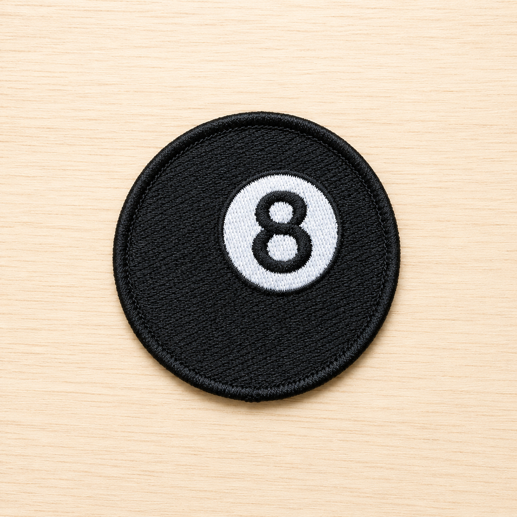 Round embroidered black magic 8 ball iron-on patch with a white circle and black number 8 design on a light wood background.