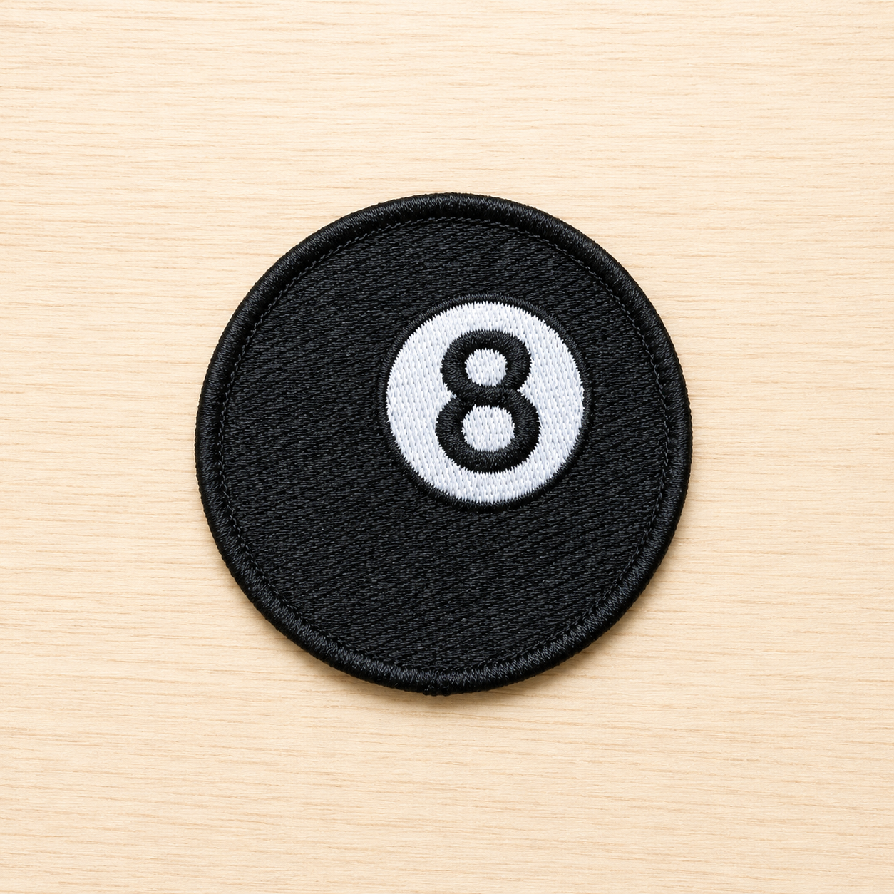 Round embroidered black magic 8 ball iron-on patch with a white circle and black number 8 design on a light wood background.
