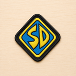Diamond-shaped embroidered SD-inspired iron-on patch with blue and yellow lettering, black border, and retro cartoon-style design on a light wood background.