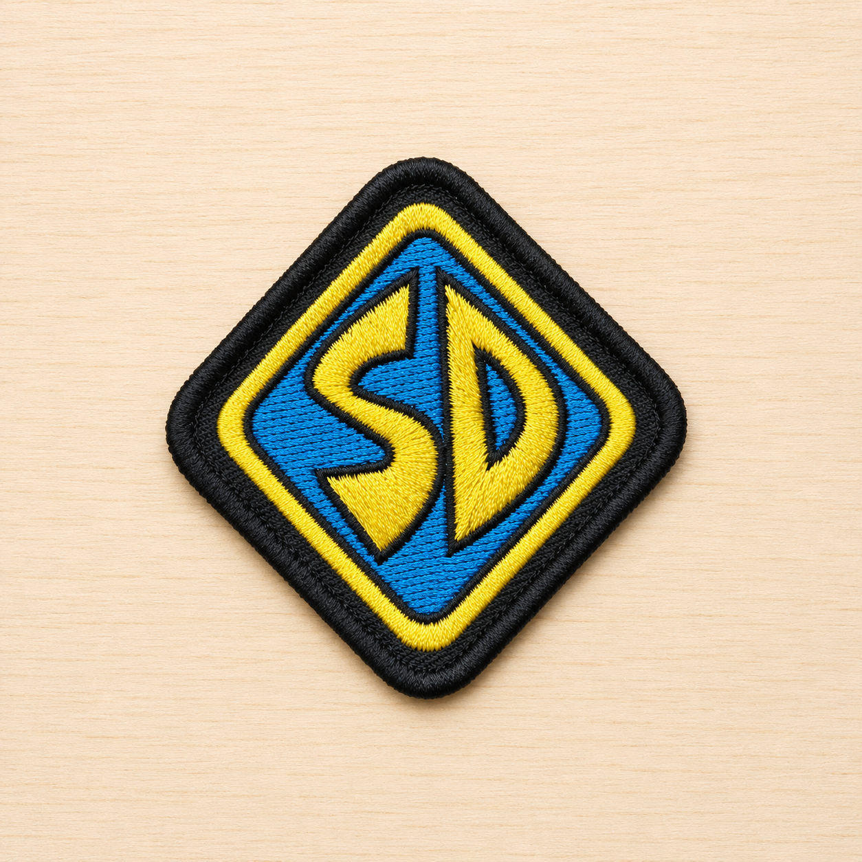 Diamond-shaped embroidered SD-inspired iron-on patch with blue and yellow lettering, black border, and retro cartoon-style design on a light wood background.