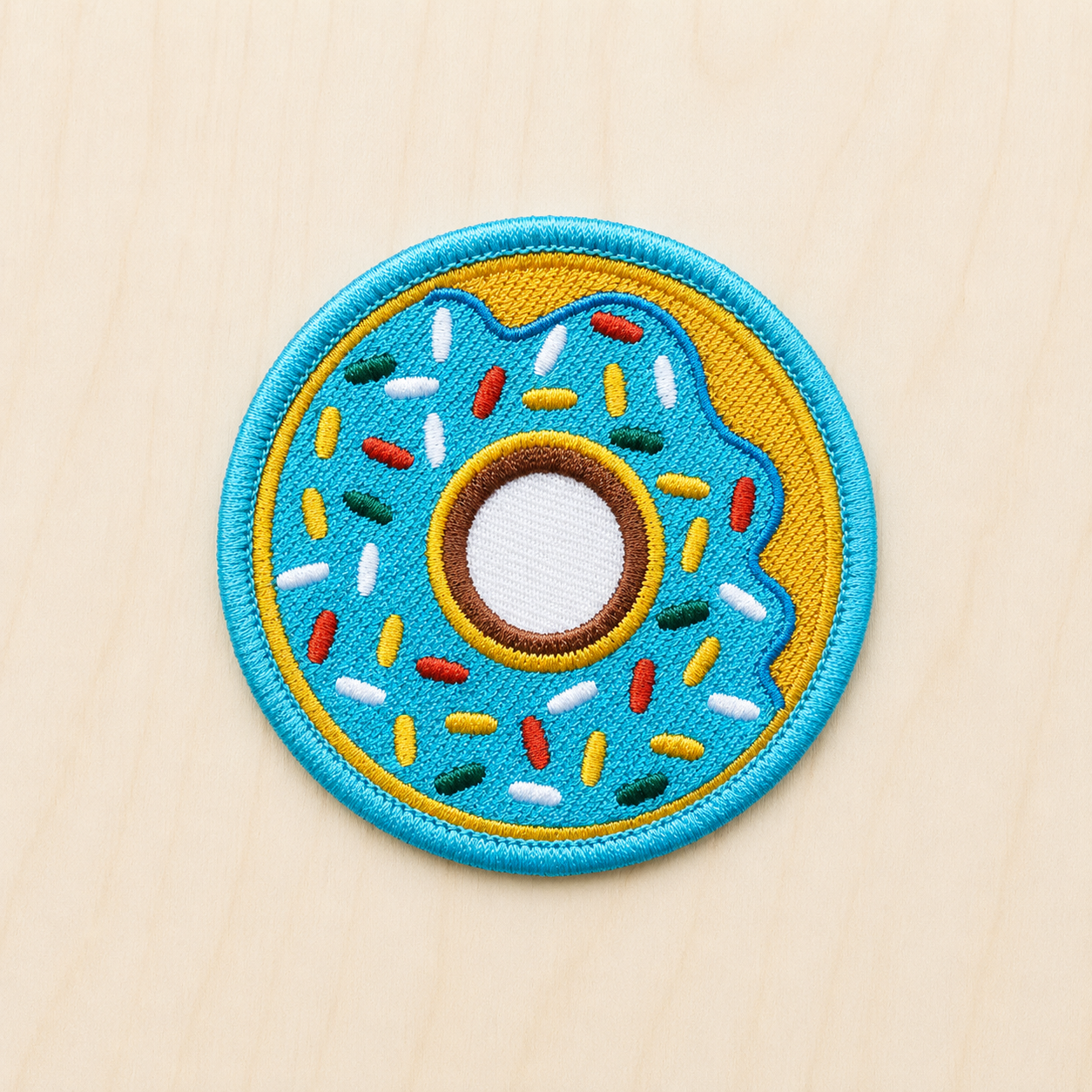 Colorful embroidered iron-on donut patch with blue frosting and rainbow sprinkles outlined in turquoise and yellow on a light wood background.