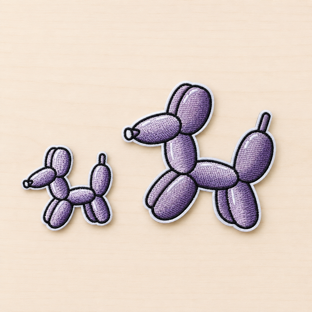 Set of two embroidered purple balloon dog iron-on patches in small and large sizes with black and white stitched borders on a light wood background.