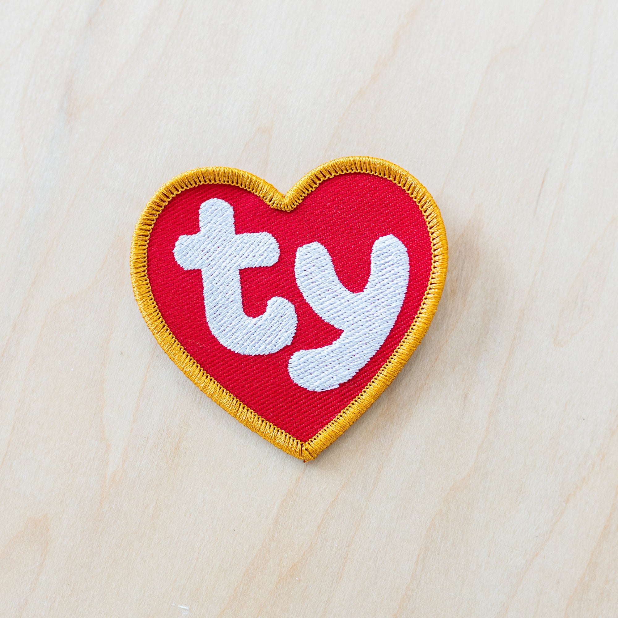 Heart-shaped embroidered iron-on patch inspired by vintage TY collectible tags, featuring bold white “ty” lettering on a bright red heart with a golden yellow stitched border on a light wood background.