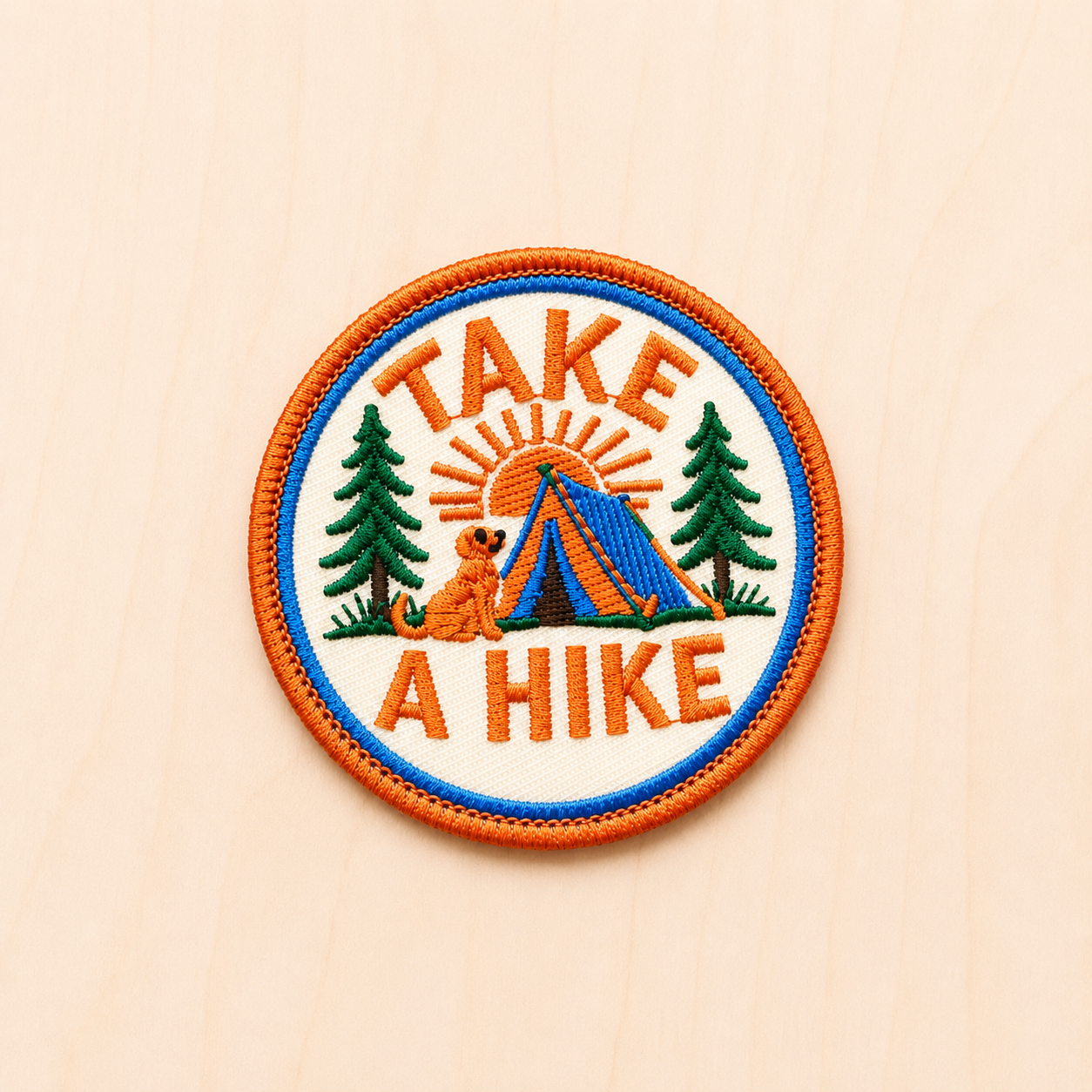 Round embroidered camping-themed iron-on patch featuring “Take A Hike” text with a tent, pine trees, sunrise, and small dog illustration in orange, blue, and green tones on a light wood background.