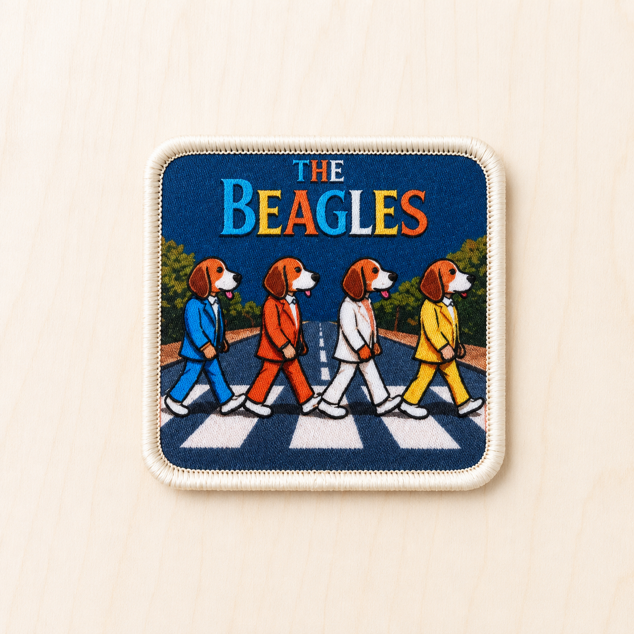Square embroidered patch inspired by a classic crosswalk album cover, featuring four beagle dogs in colorful suits walking across a striped road against a dark blue background with “The Beagles” text at the top, displayed on a light wood surface.
