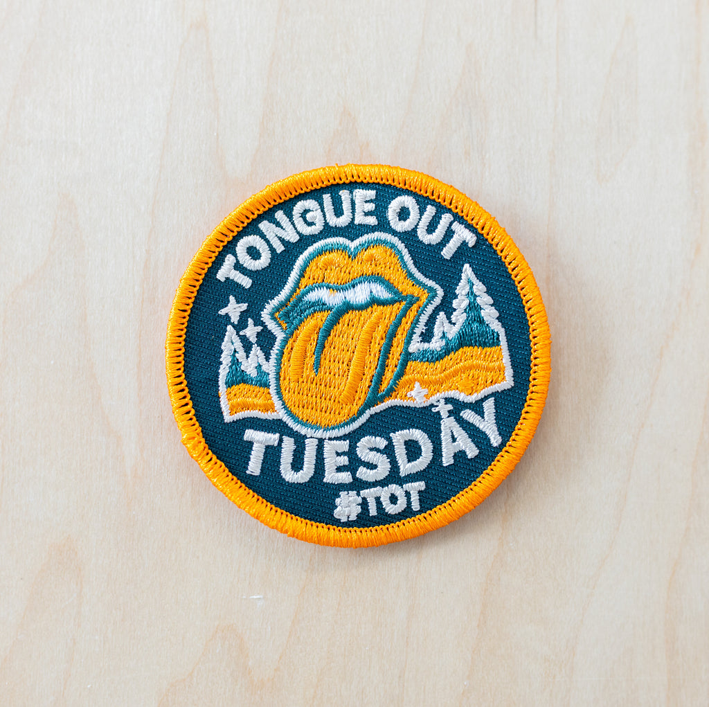 Round embroidered iron-on patch featuring a retro tongue-and-lips design with snowy mountains, pine trees, and “TONGUE OUT TUESDAY #TOT” text in teal, orange, and cream colors on a light wood background.