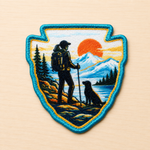 Arrowhead shaped embroidered iron-on patch featuring a hiker with trekking poles and a dog overlooking a mountain lake at sunset, with pine trees and snow-capped peaks in blue, orange, and teal tones on a light wood background.