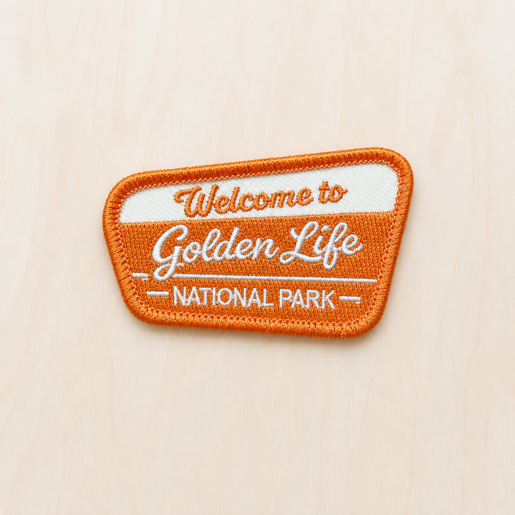 Orange and cream embroidered iron-on patch reading “Welcome to Golden Life National Park” displayed on a light wood background in a retro national park style.
