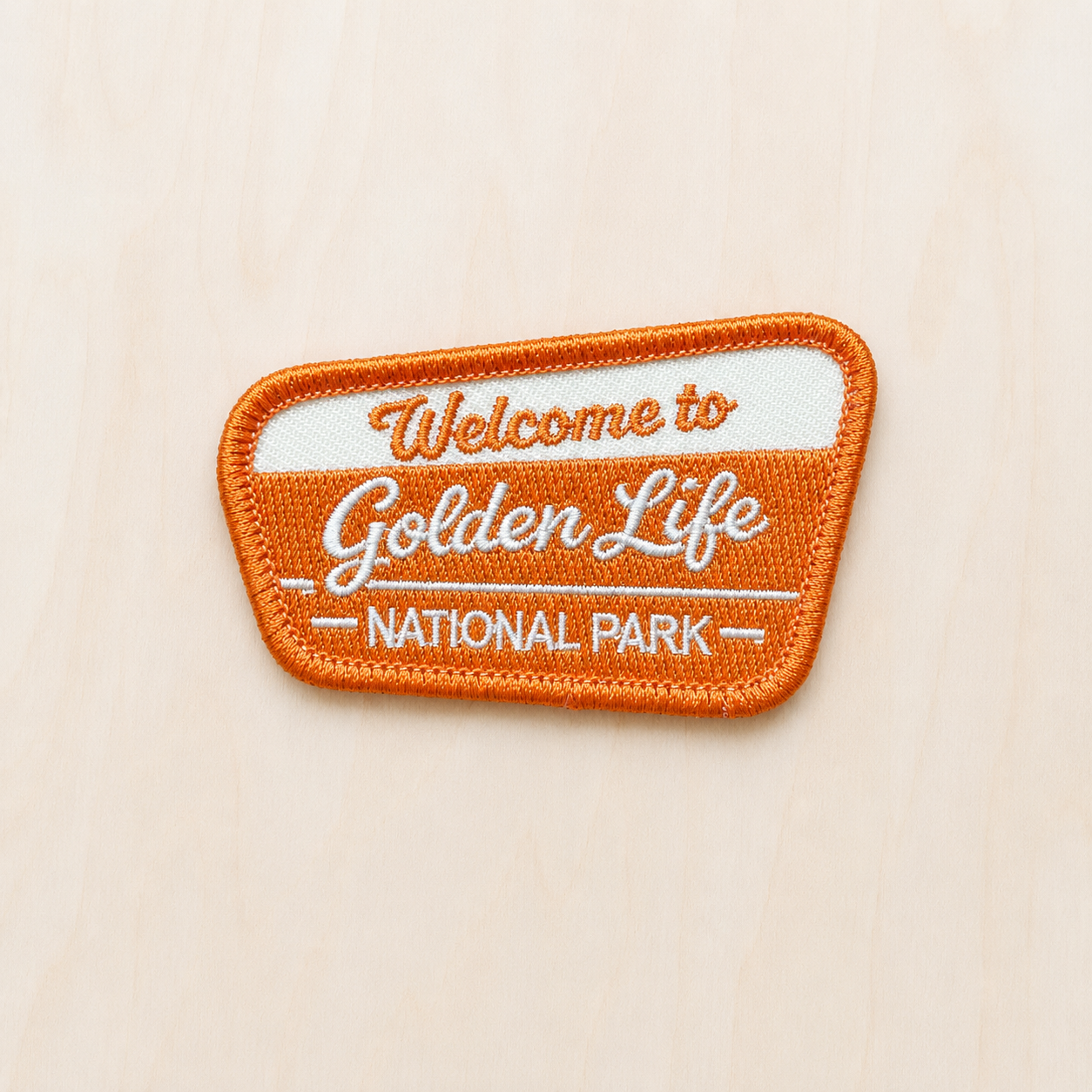 Orange and cream embroidered iron-on patch reading “Welcome to Golden Life National Park” displayed on a light wood background in a retro national park style.