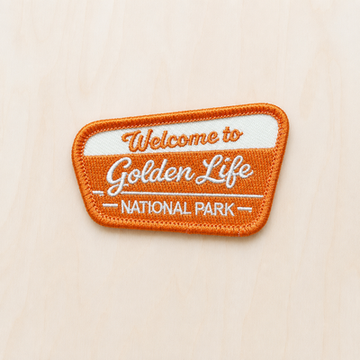 Orange and cream embroidered iron-on patch reading “Welcome to Golden Life National Park” displayed on a light wood background in a retro national park style.