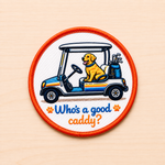 Round embroidered golf-themed iron-on patch featuring a golden retriever riding in a golf cart with golf clubs and “Who’s a good caddy?” text in blue and orange lettering on a light wood background.