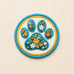 Circular embroidered iron-on patch featuring a floral paw print filled with yellow, white, and blue wildflowers outlined in turquoise and gold on a light wood background.