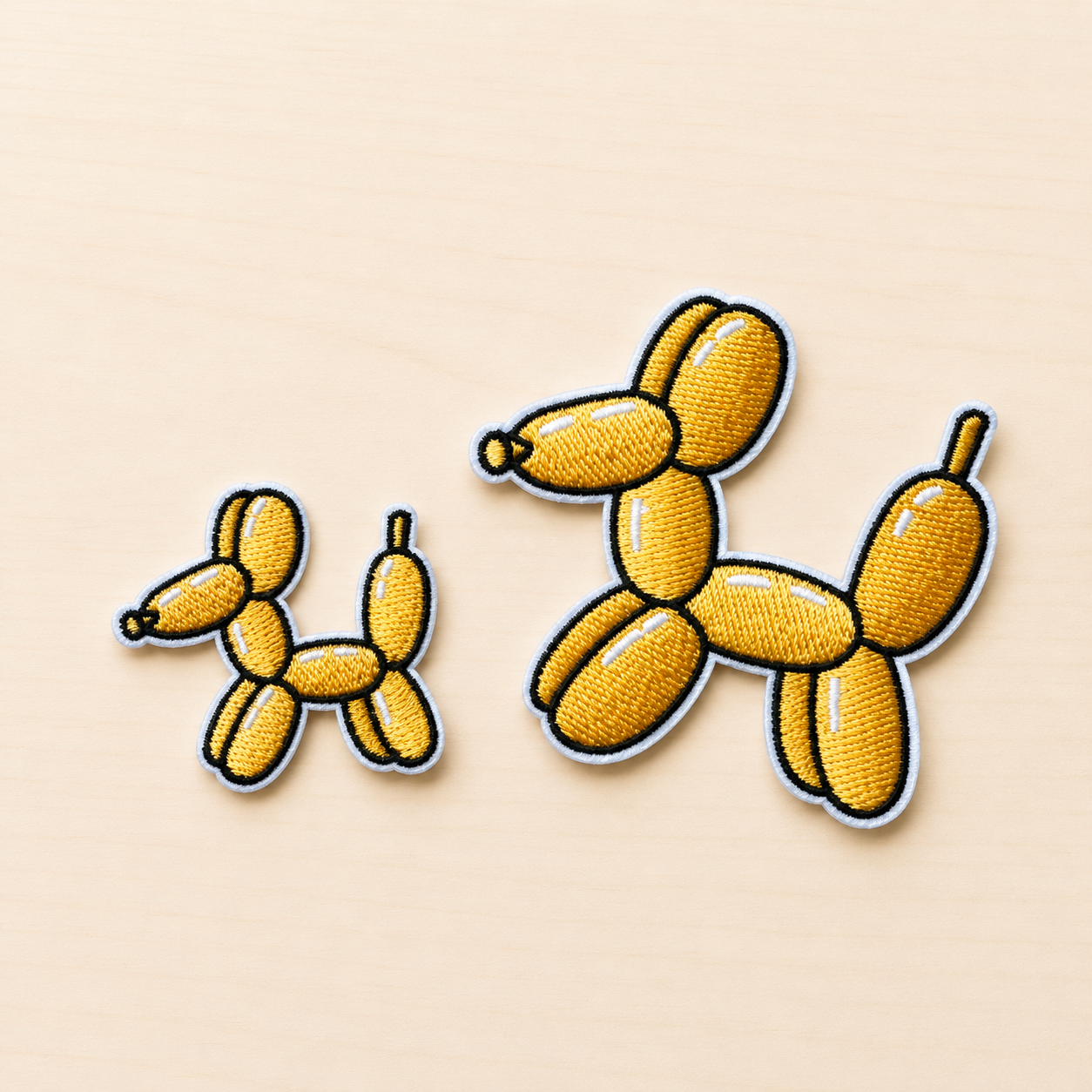 Set of two embroidered yellow balloon dog iron-on patches in small and large sizes with black and white stitched borders on a light wood background.