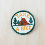 Round embroidered patch featuring a camping tent, pine trees, and the phrase “Take a Hike” on a light background