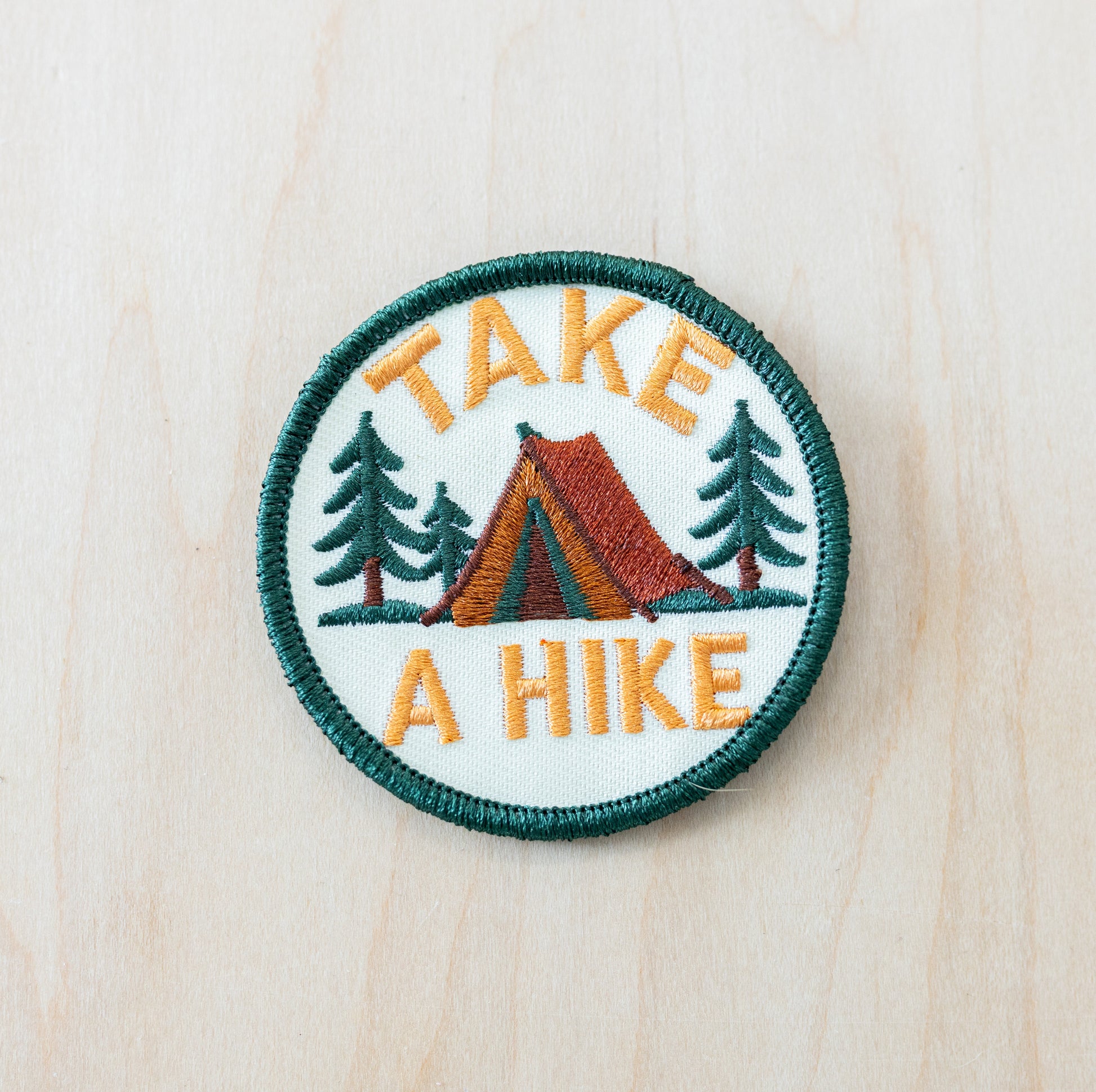 Round embroidered patch featuring a camping tent, pine trees, and the phrase “Take a Hike” on a light background