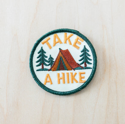 Round embroidered patch featuring a camping tent, pine trees, and the phrase “Take a Hike” on a light background